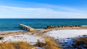 A beach with a wooden pier extending into the sea, a rocky breakwater, and sparse vegetation. - Lovely holiday home near beach with sauna (Otterup)