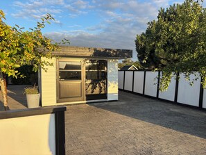 Un espace extérieur de taille réduite, comprenant une petite construction, peut-être un abri de jardin, jouxtant un mur blanc bordé d’une bande noire.