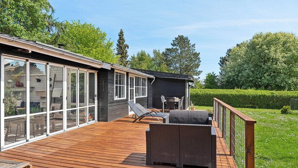 Um deck de madeira com mobiliário externo, uma sala envidraçada e vista para uma paisagem verde.
