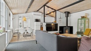 Uma sala de estar moderna com um sofá cinzento, uma área de jantar com cadeiras brancas, uma televisão, uma lareira e um armário verde.