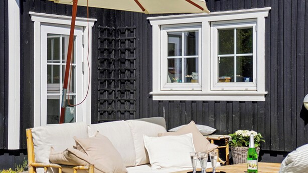A black wooden building with white trim, a white-framed window, a wicker sofa, a wooden table with glasses and a bottle, and a wicker chair. - Bright and spacious holidayhome near Vejby Strand (Vejby)