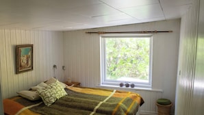 Ein Schlafzimmer mit Bett, Fenster mit Blick ins Grüne, einem Bild an der Wand und einem Nachttisch mit Lampe.