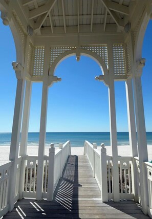Een houten terras dat leidt naar een strand, met een witte gazebo-constructie.