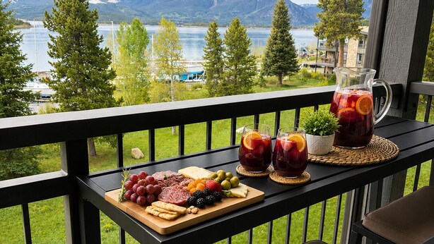 A balcony with a view of mountains and a lake, a wooden board with various cheeses and fruits, and two glasses of red drink. - Dillon Lakefront Luxury w Iconic Mountain Views. Walk to Marina & Amphitheater (Dillon)