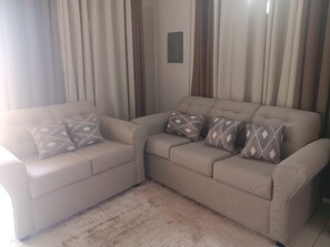 Ein Wohnzimmer mit einem beigefarbenen Sofagarnitur, gemusterten Kissen und einem hellen Teppich.
