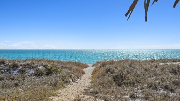 Un sentier de sable bordant la plage, qui mène à une mer d’un bleu limpide, avec une végétation clairsemée.
