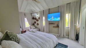 A bedroom with a large bed, a flat-screen TV mounted on the wall, and a window with curtains.