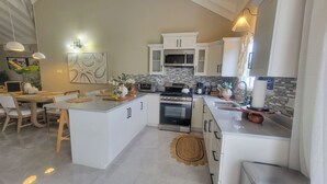 A modern kitchen with white cabinetry, a central island, a microwave, an oven, and a sink. There are pendant lights, a vase with flowers, and a painting on the wall.
