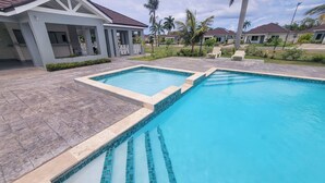 A swimming pool with tiled edges, surrounded by a paved patio and a covered area with chairs.