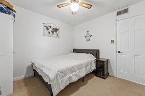 Una habitació amb un llit, una tauleta de nit, un mapa del món, una decoració penjada, un ventilador de sostre i una porta blanca.