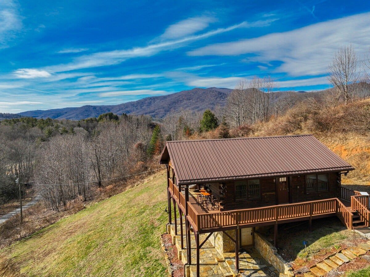 A cabin with a wooden deck and a stone foundation, surrounded by a hilly landscape.