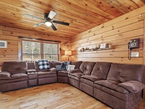Ein gemütliches Blockhaus-Interieur mit einem braunen Leder-Modulsessel, Deckenventilator, Holzwänden und einem Fenster, das für natürliches Licht sorgt.
