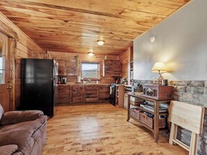 Ein rustikales Blockhäuschen im Inneren mit Holzbalkendecke, einer Küchenzeile mit Kühlschrank, Herd und Holzausbau sowie einer Sitzecke mit einer Couch.