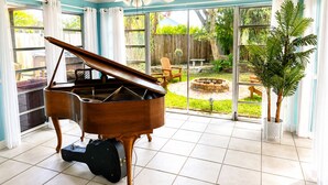 Amplia habitación con un piano de cola, ventilador de techo y grandes ventanas que dan a un patio trasero con una chimenea.