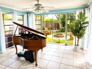 Amplia habitación con un piano de cola, ventilador de techo y grandes ventanas que dan a un patio trasero con una chimenea.