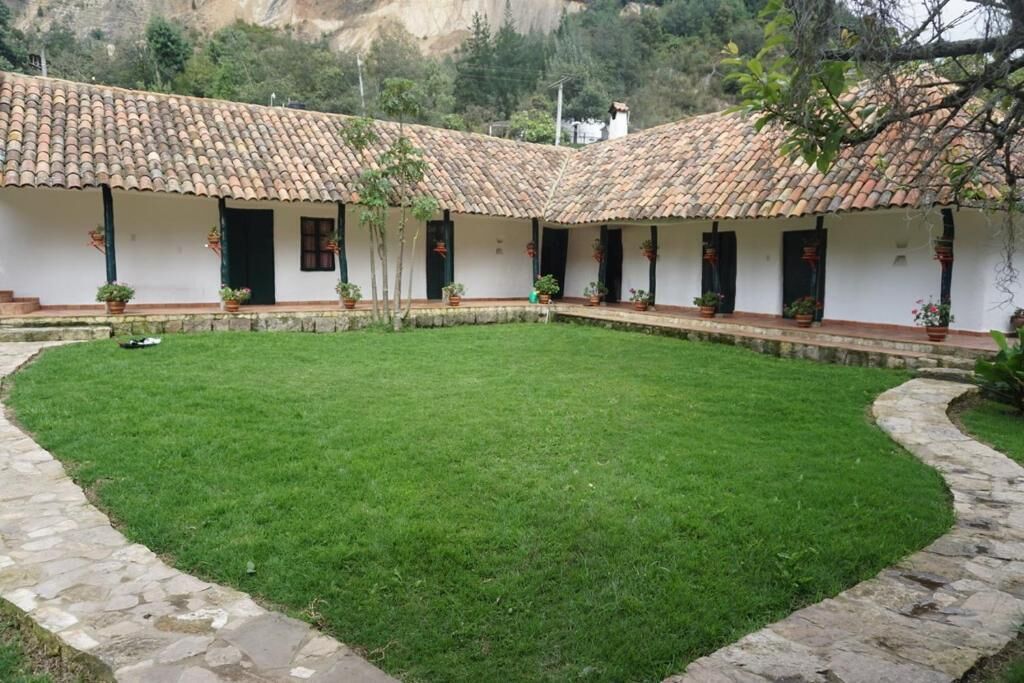 A courtyard with a white building featuring a tiled roof and multiple windows, surrounded by a stone pathway and green lawn.