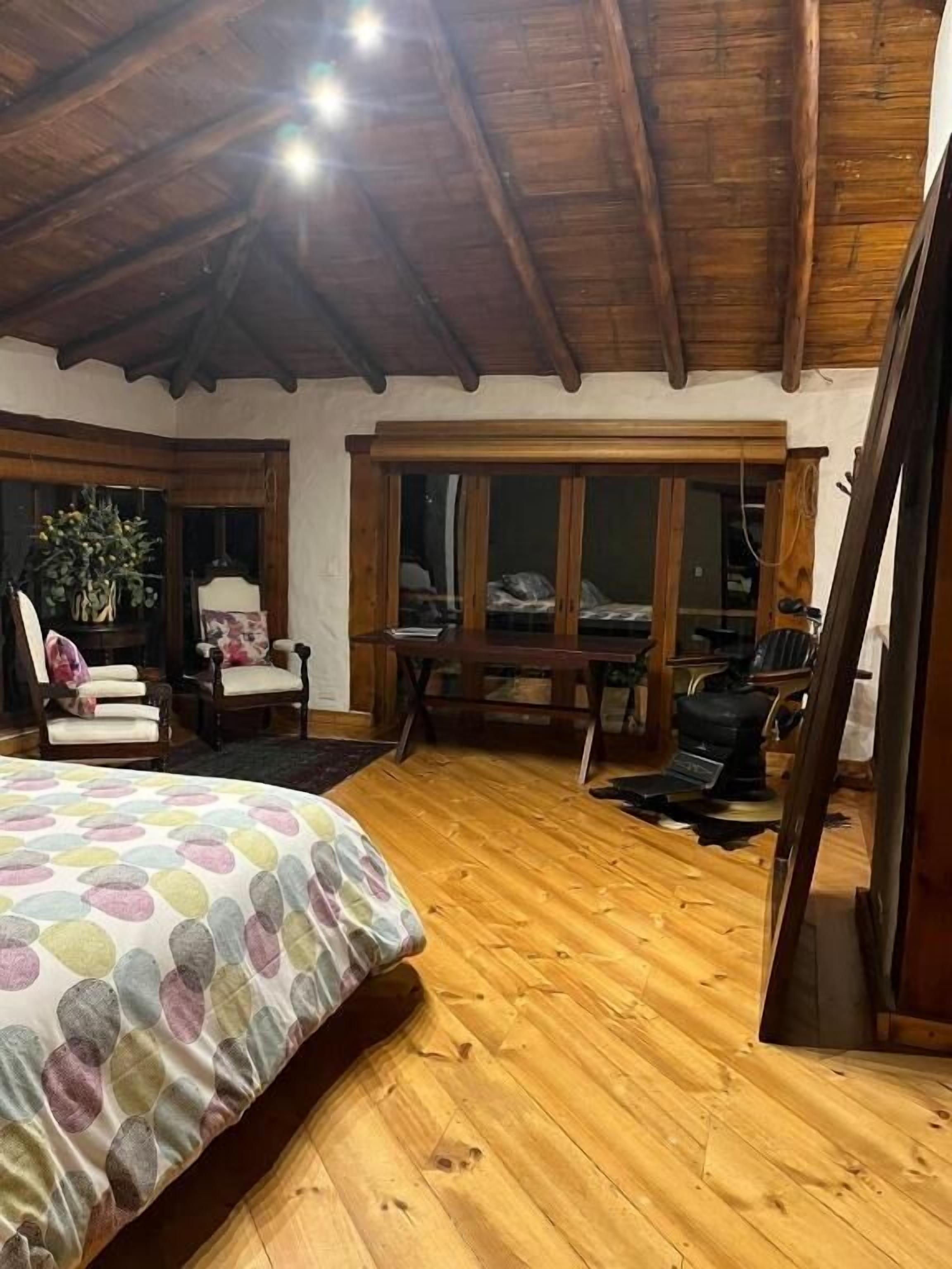 Um quarto espaçoso com piso de madeira, uma cama grande com colcha colorida, teto de madeira e uma janela ampla com vários painéis.