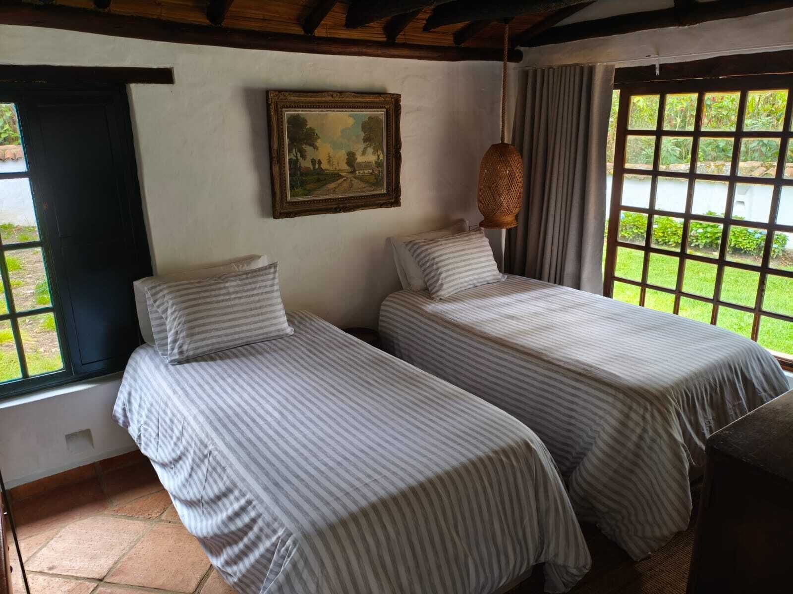 A room with two beds, a painting on the wall, and a window with a view of greenery.