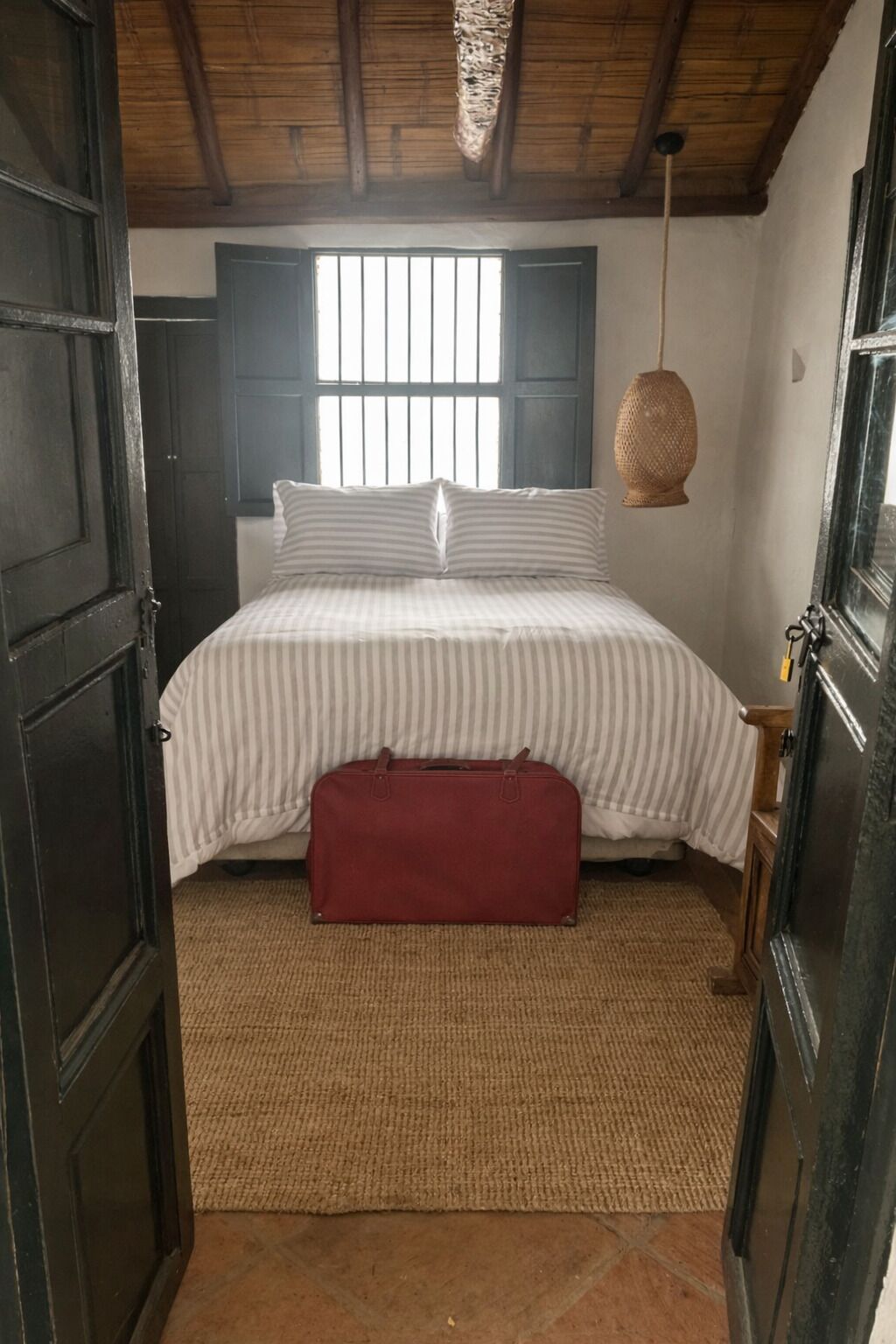 Ein Schlafzimmer mit einem Bett, einem roten Koffer, einem Teppich und einem Fenster mit Gitterstäben.
