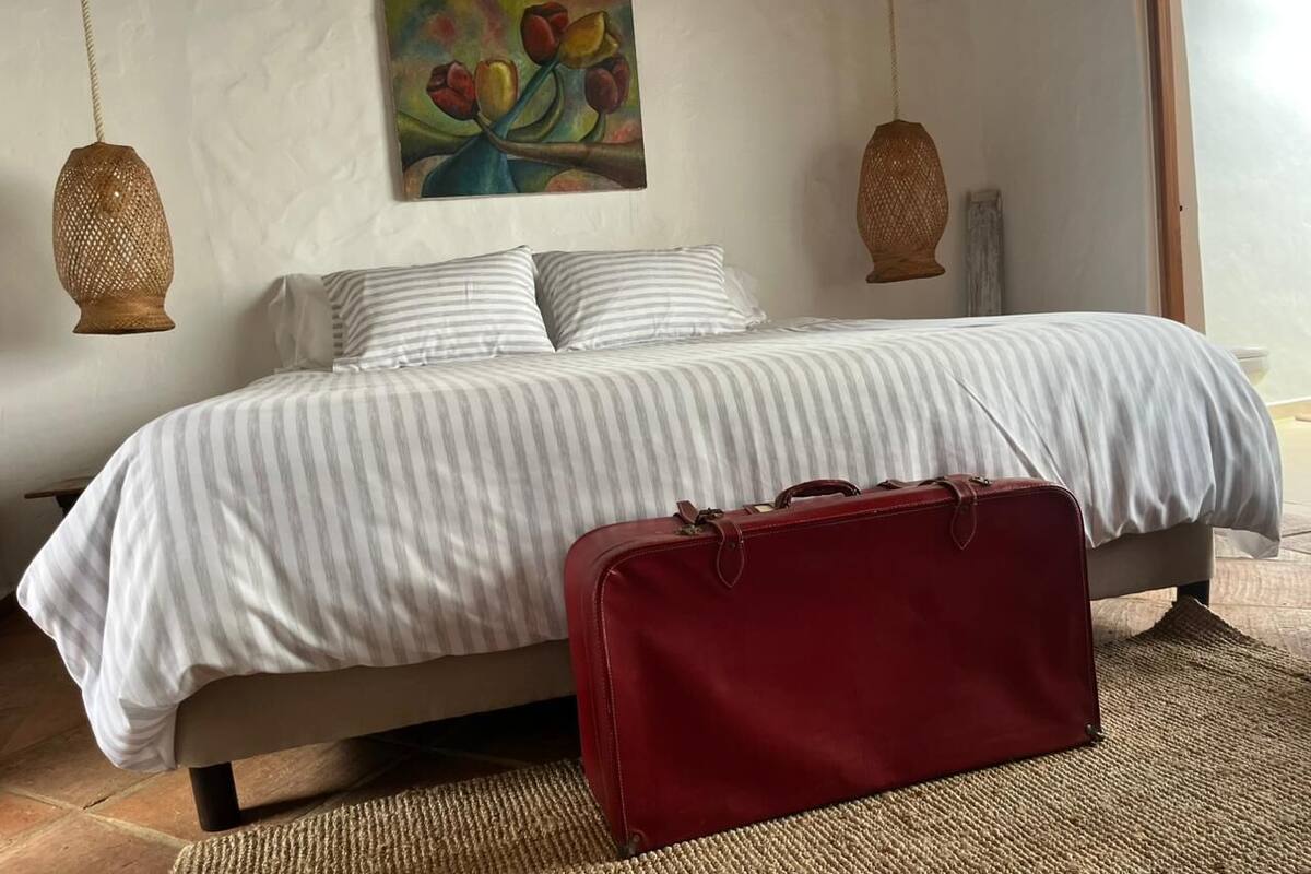 Un dormitorio con cama, ropa de cama a rayas, una maleta roja y dos lámparas colgantes.