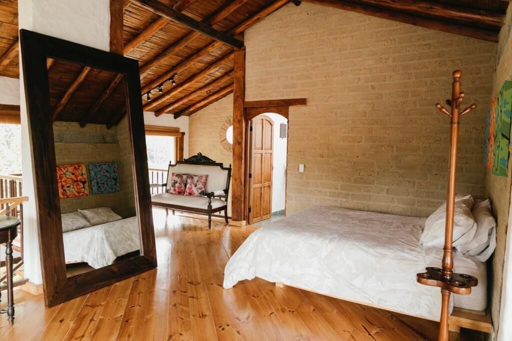 Un dormitorio rústico con techo de madera, un espejo grande, una cama con ropa de cama blanca y un perchero de madera.