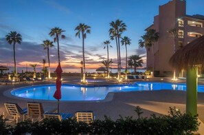Un área de piscina con sillones, palmeras y un edificio con balcones.