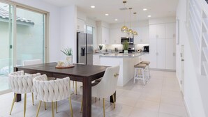 Una cocina moderna con armarios blancos, una isla central, electrodomésticos de acero inoxidable y un comedor con mesa de madera oscura y sillas tapizadas en blanco.