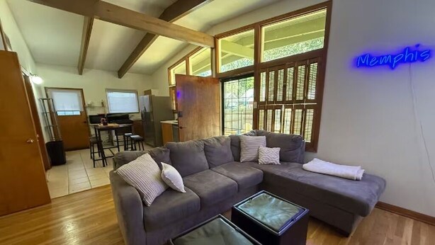 A modern living room with a sectional sofa, two ottomans, a kitchen area in the background, and a neon sign reading "Memphis". - Golf and Campus Getaway  EV, Near Hospitals- 3 Bedroom, 2.5 Bath (Memphis)