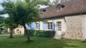 Ein Gebäude aus Stein mit einem Ziegeldach, ein Baum im Vordergrund und ein Hecke entlang der Seite.
