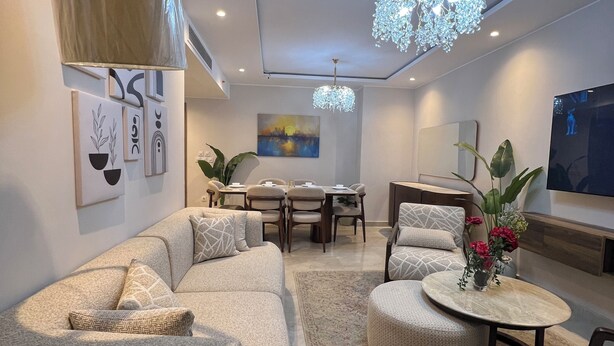 A modern living room with a sectional sofa, a round coffee table, a dining area with a table and chairs, and a chandelier.