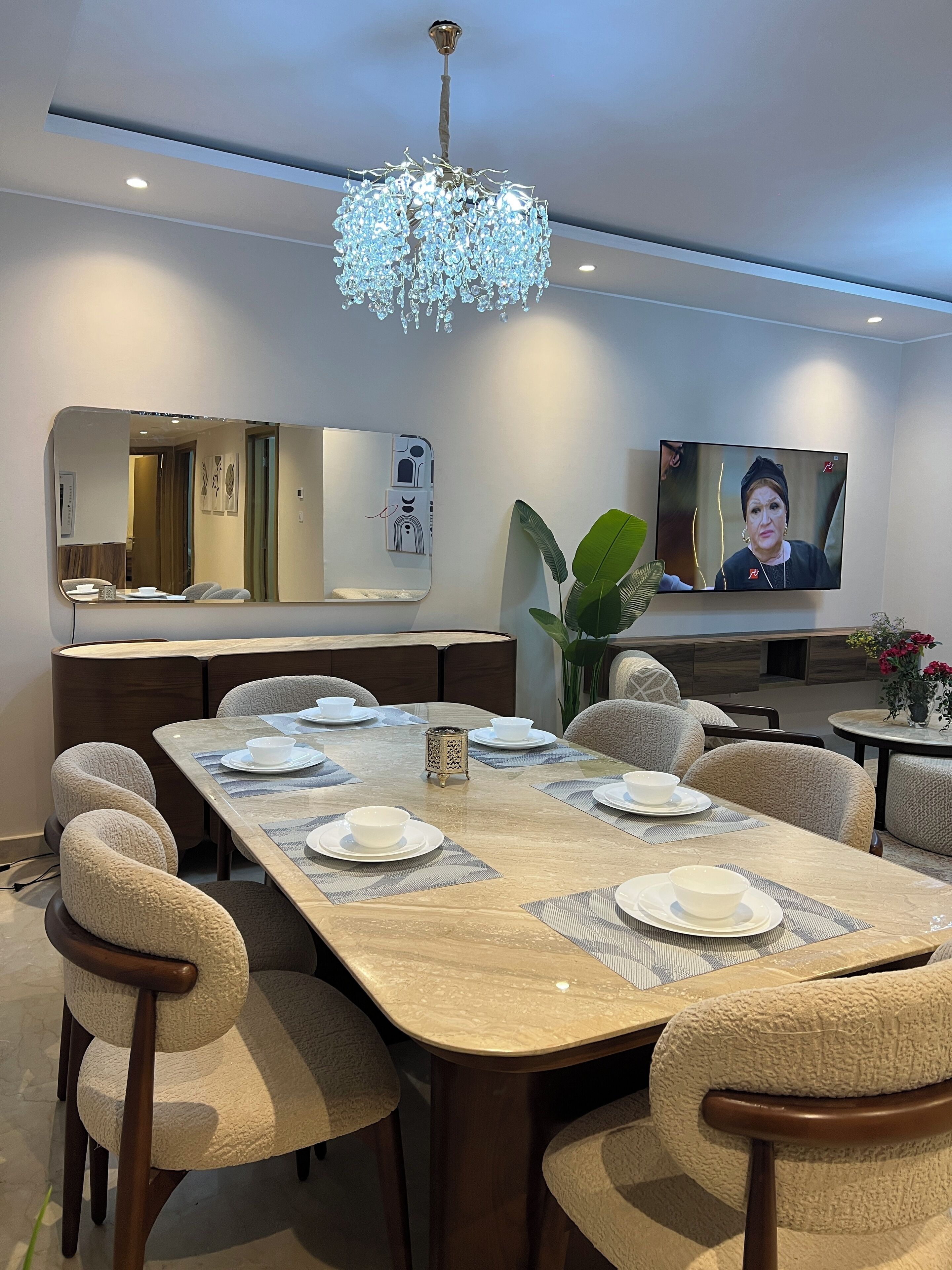 Un coin repas avec une longue table dressée pour quatre personnes, des chaises recouvertes d’un tissu texturé, un lustre, un téléviseur et un miroir orné d’un motif représentant une coque de téléphone.