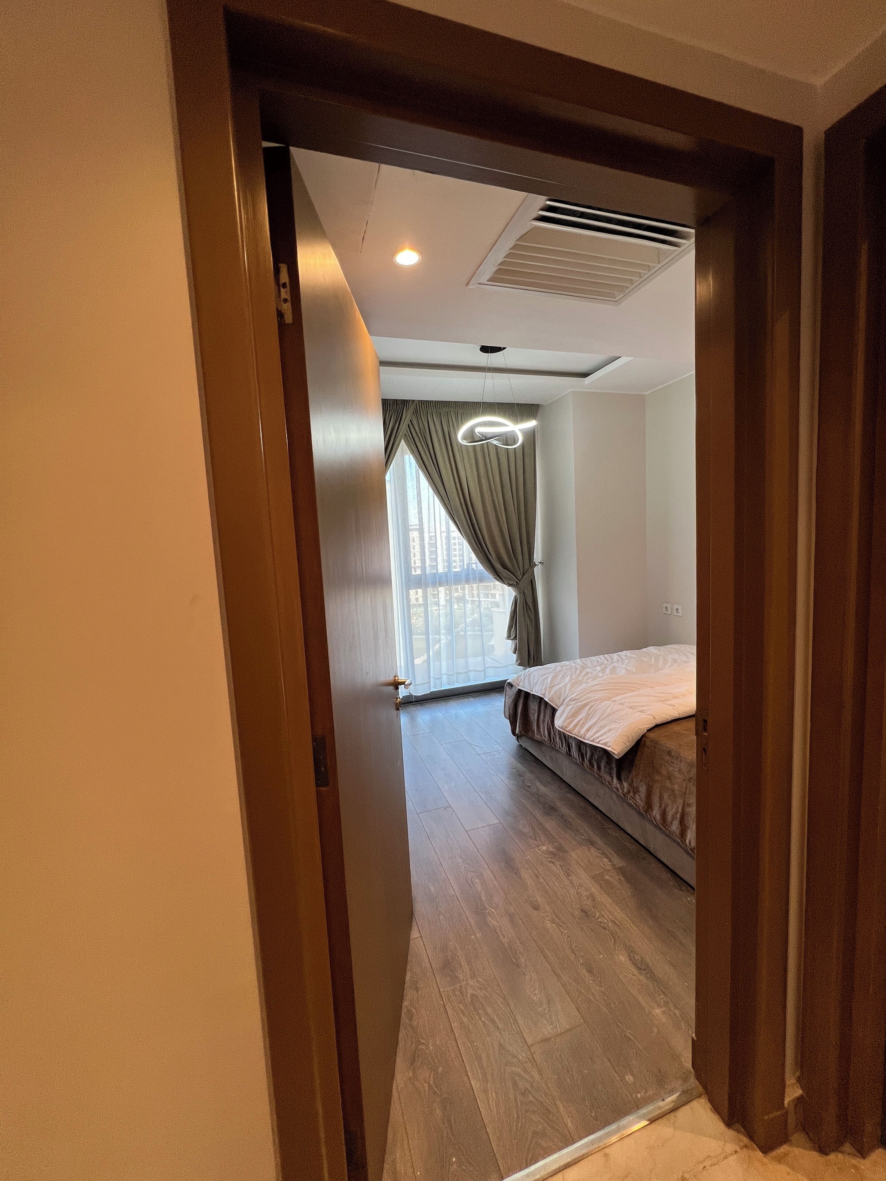 Una habitación de hotel con cama, ventana con cortinas, lámpara de techo y puerta.