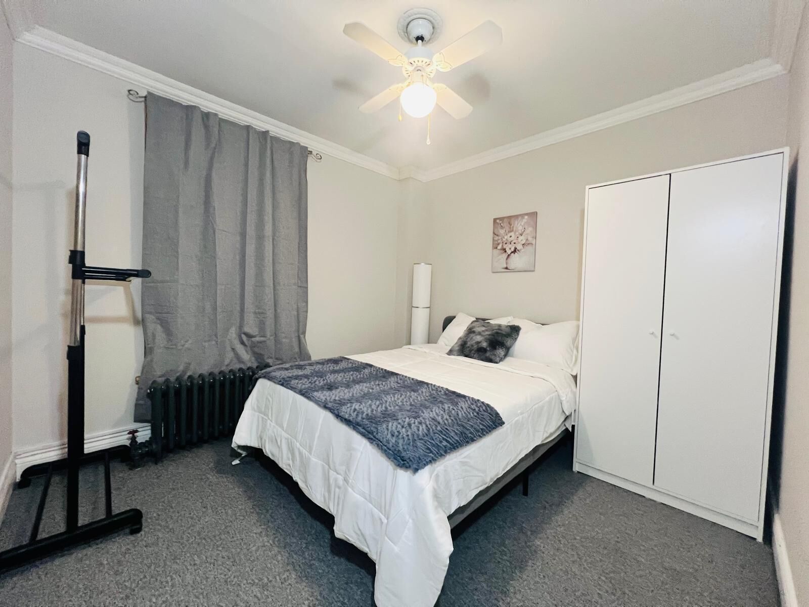 Un dormitorio con cama, radiador, ventilador de techo, armario y un cuadro en la pared.