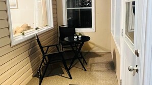 Een smalle gang met een zwarte klapstoel en een kleine tafel met twee witte kannen. Er zijn twee ramen, waarvan er één uitzicht geeft op een huis en de ander een decoratief scherm heeft.