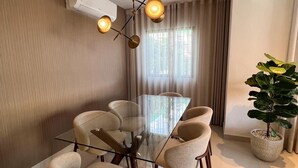 A modern dining room with a glass table and six chairs, a pendant light, a potted plant, and a large window with curtains. - Quiet and cozy place (Santo Domingo)