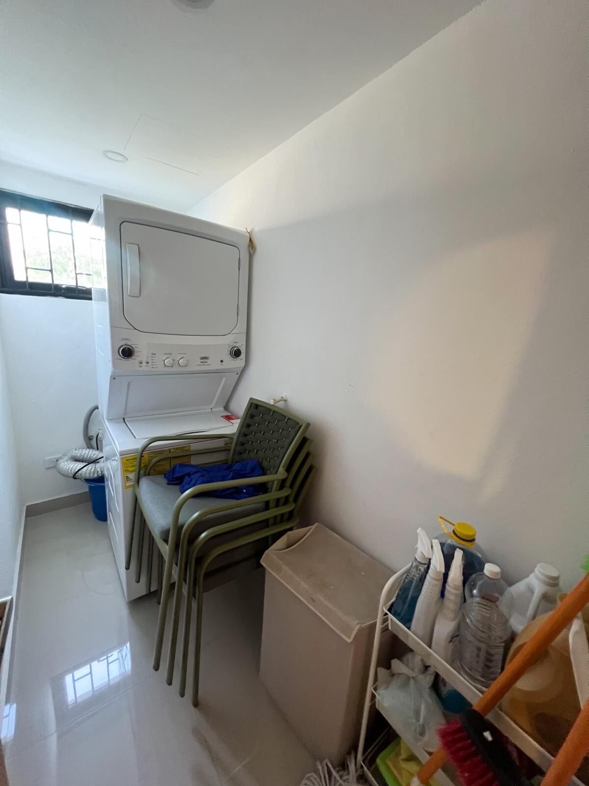 Une petite pièce comprenant un lit, une petite table, une chaise, une poubelle et une étagère avec des bouteilles.