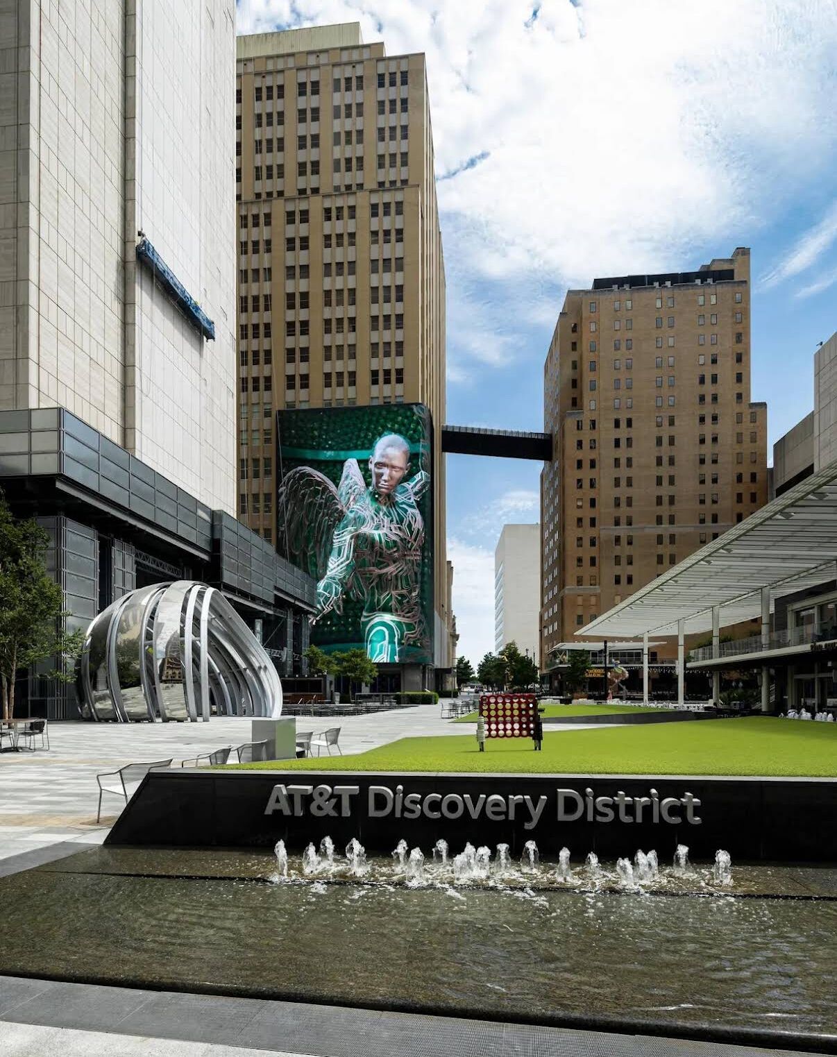 A modern urban plaza with a large digital billboard, a water feature, and the AT&T Discovery District sign.