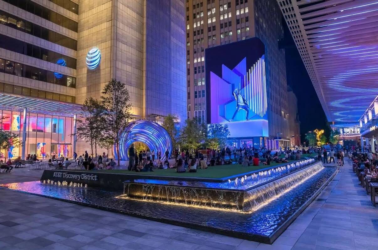 A public square with a water feature, AT&T Discovery District signage, and a large digital display.