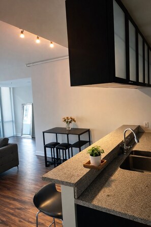 A modern kitchen with a granite countertop, a black chair, a small table with a vase of flowers, and a large window. - Bright High-Rise 1BD Prime Downtown Stay (Dallas)