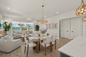 A spacious living area with a glass dining table, white furniture, and a view of the ocean. - Sun & Salt Retreat: 5BR/4.5BA NEW Pool Home! (Fort Myers Beach)