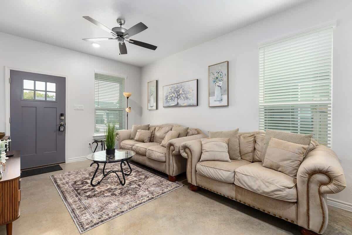 A living room with a beige sofa, a coffee table, and a ceiling fan. There are framed pictures on the wall and a door with a window.