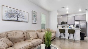 Ein modernes Wohnzimmer mit einem beigefarbenen Sofa, einem Glastisch und einer Topfpflanze. Angrenzend befindet sich eine Küche mit grauen Schränken, einer Mikrowelle und einem Kühlschrank.
