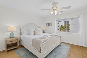 A bedroom with a white bed, a nightstand with a lamp, a ceiling fan, and a window with a view of palm trees. - New Listing! Mariners Walk 3F- Ocean View Balcony! (Isle of Palms)