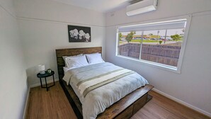 Una camera da letto con testiera in legno, un'ampia finestra vista parcheggio, un comodino con una lampada e un condizionatore d'aria a parete.