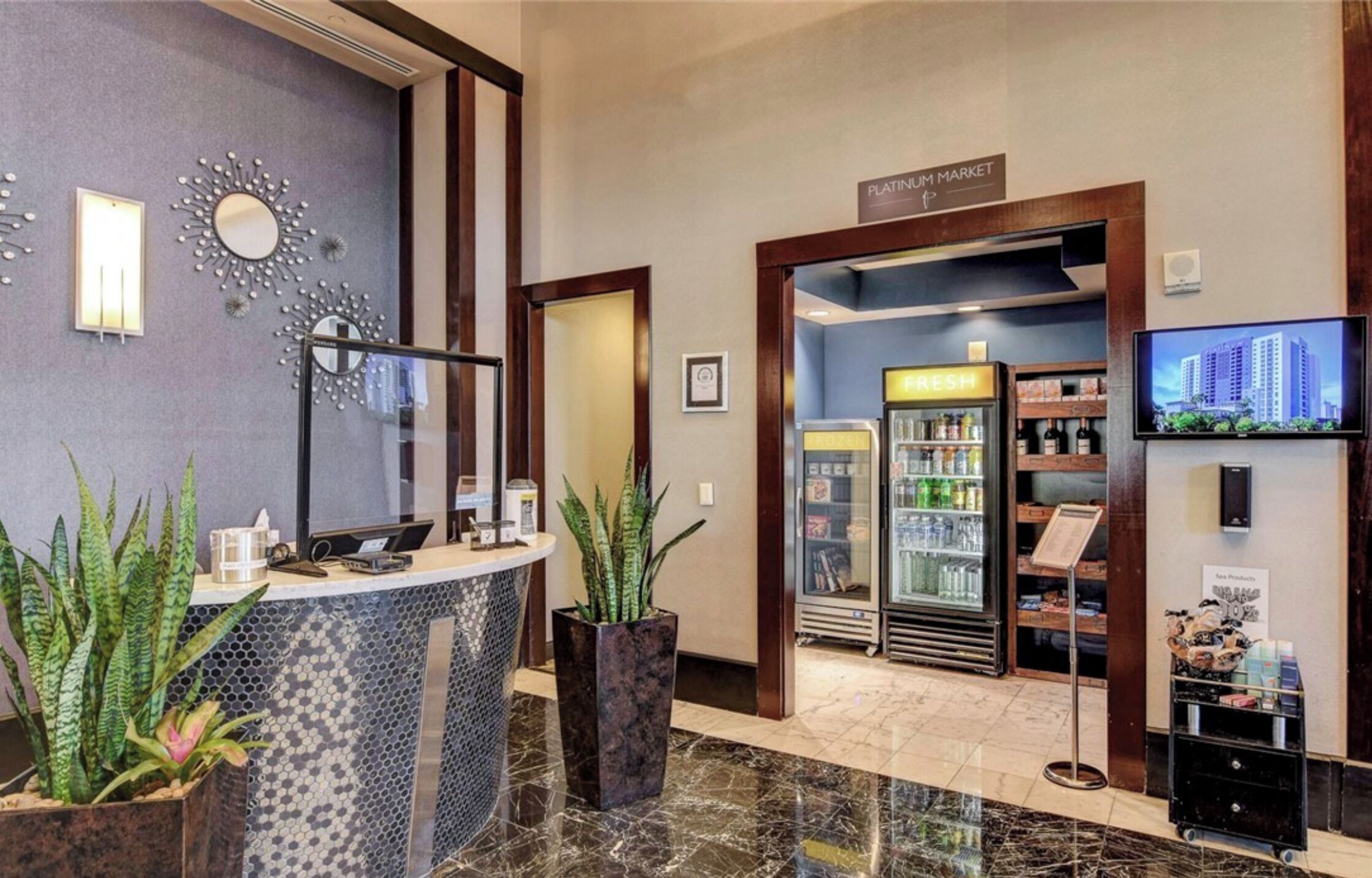 A hotel lobby with a reception desk, a Platinum Market sign, a refrigerator with beverages, and a television displaying a cityscape.