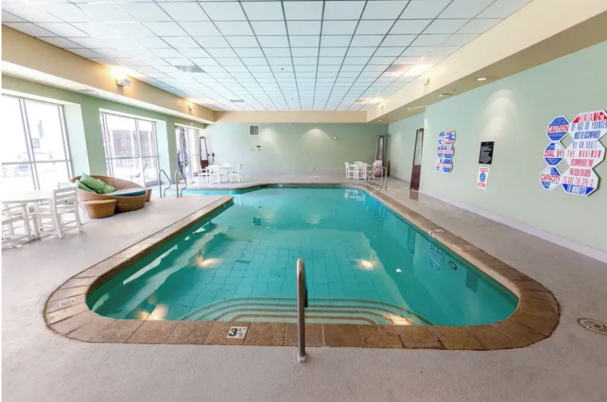 An indoor swimming pool with a diving board, lounge chairs, and safety signs on the walls.