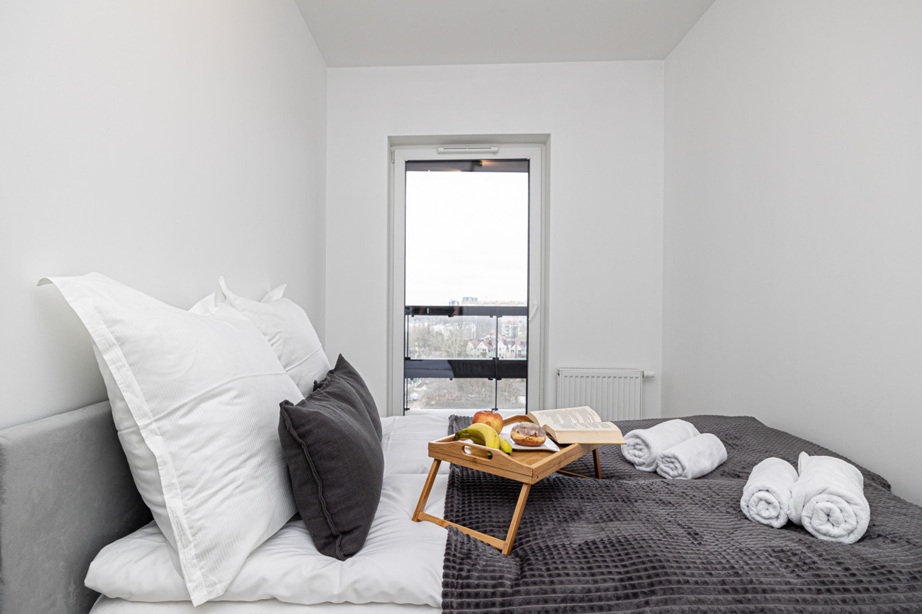 Hotellihuone, jossa on sänky, tyynyjä, hedelmiä sisältävä tarjotinalusta ja kirja sekä ikkuna, josta on näkymä.