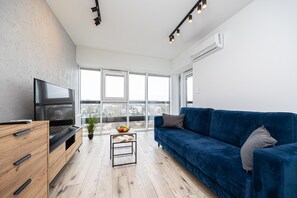 Ein modernes Wohnzimmer mit Fernseher, einem blauen Sofa, einem Holzkasten und einem kleinen Tisch mit Pflanze und Obstschale.