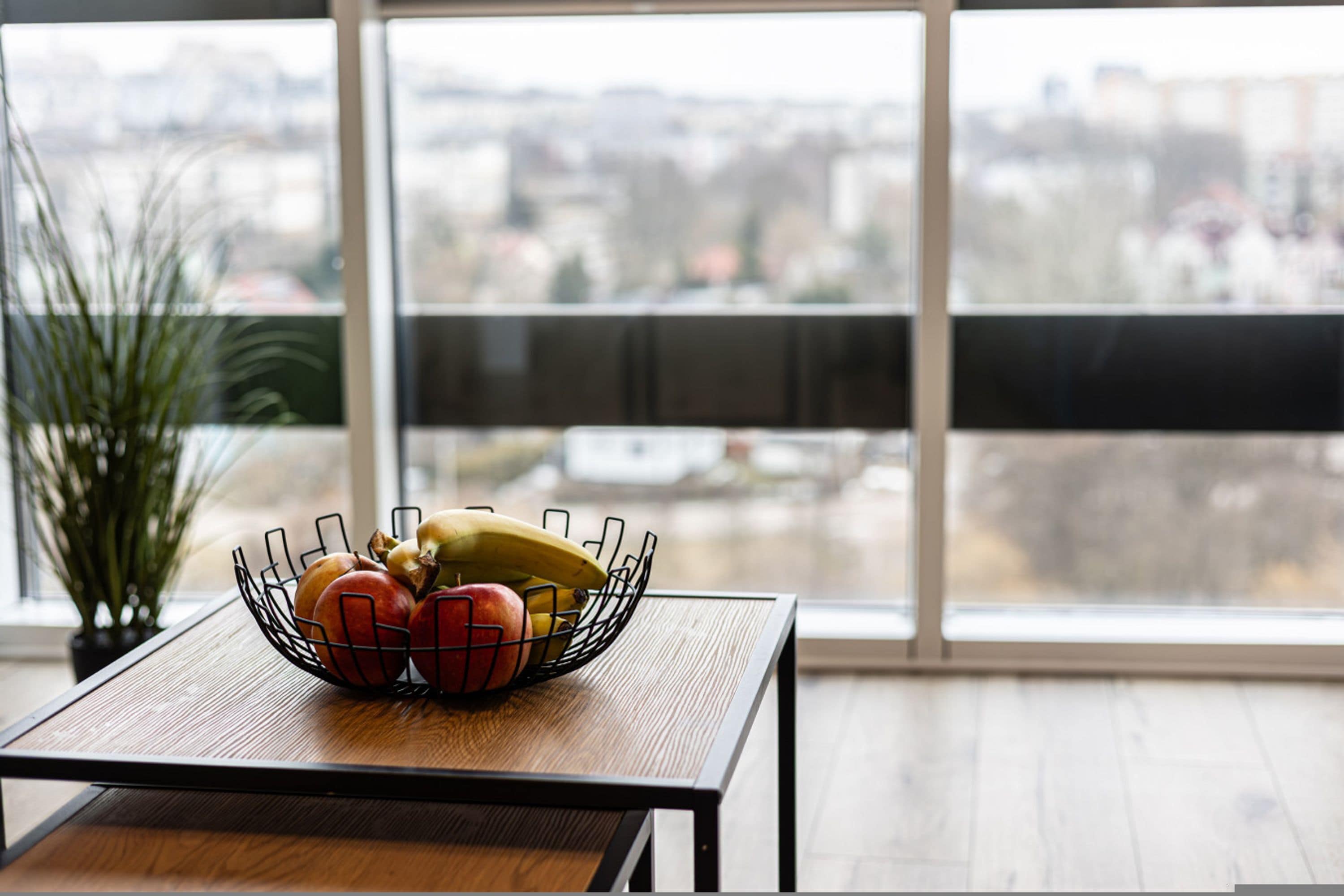 Une table à manger sur laquelle se trouve un saladier rempli de pommes et de bananes, une plante en pot et, à travers de grandes fenêtres, une vue sur le paysage urbain.