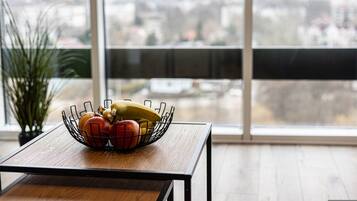 Une table à manger sur laquelle se trouve un saladier rempli de pommes et de bananes, une plante en pot et, à travers de grandes fenêtres, une vue sur le paysage urbain.